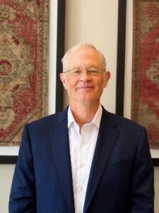 Brad Smiling older man in a navy suit and white shirt stands in front of framed patterned rugs.