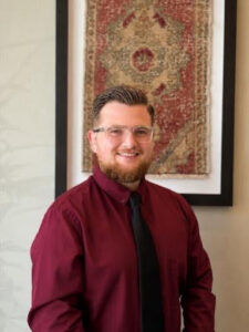 Braxton Man in a burgundy dress shirt and black tie, smiling at the camera with framed rug artwork behind him.