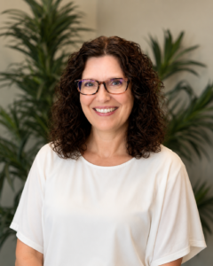 Brenda Smiling woman with curly brown hair, glasses, and a white blouse, standing in front of leafy plants.