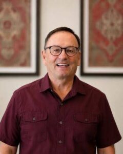 Eric Smiling man with glasses in a maroon short-sleeved shirt poses for a portrait in front of framed art behind him.