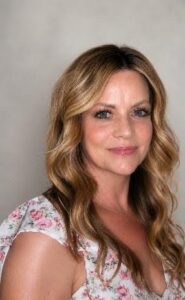 Holly Portrait of a woman with long wavy blonde hair, wearing a light floral top, facing camera with a gentle smile against a gray backdrop.