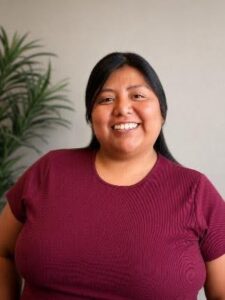 Ivette Smiling woman in a burgundy knit shirt standing indoors with a plant in the background.