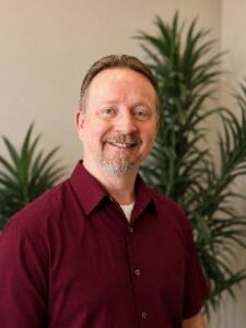Jared Smiling man in a burgundy button-down shirt standing indoors with tall green plants in the background.