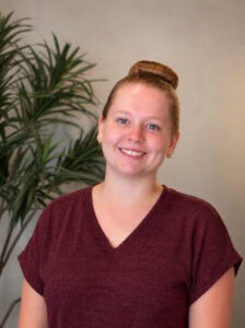 Mikayla Smiling woman with a bun wearing a burgundy V-neck top, indoors with a leafy plant behind her.