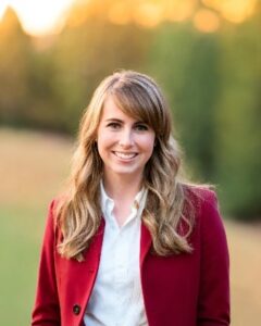Mindy Portrait of a smiling woman with wavy blonde hair, wearing a white shirt and red blazer, outdoors in a sunlit park.