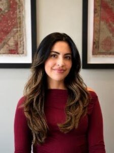 Yasmeen Portrait of a woman with long wavy dark hair wearing a burgundy top, smiling in a light, neutral office setting with framed art behind her.